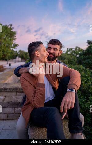 Affectueux heureux couple gay, assis sur un mur au coucher du soleil Banque D'Images