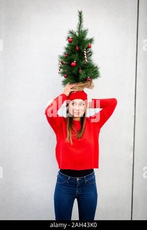 Woman wearing red pull et chapeau, wolly holding arbre de Noël artificiel sur le dessus de sa tête, en face d'un mur Banque D'Images