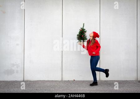 Woman wearing red pull et chapeau, wolly holding arbre de Noël artificiel devant un mur Banque D'Images