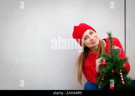 Woman wearing red pull et chapeau, wolly holding arbre de Noël artificiel devant un mur Banque D'Images