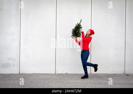 Woman wearing red pull et chapeau, wolly holding arbre de Noël artificiel devant un mur Banque D'Images