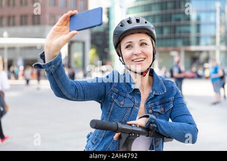 Femme souriante avec e-scooter en tenant une dans la ville selfies, Berlin, Allemagne Banque D'Images