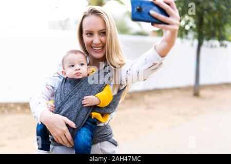 Happy mother with baby boy dans une écharpe en tenant un selfies Banque D'Images