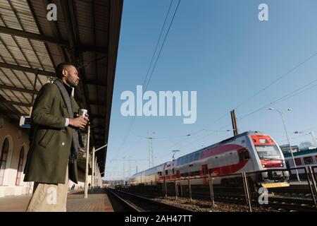 L'homme élégant avec tasse réutilisable attendant le train Banque D'Images