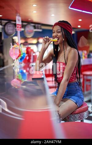 Jeune femme noire de tresses hairstyle assis sur un tabouret rouge restaurant's bar de mordre un morceau de citron Banque D'Images