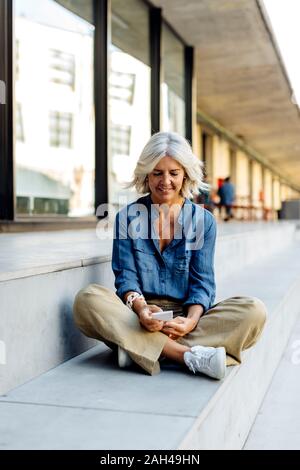 Femme d'âge mûr assis en tailleur sur des pas dans la ville, using smartphone Banque D'Images