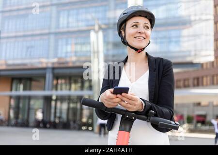 Femme heureuse avec l'e-scooter et smartphone dans la ville, Berlin, Allemagne Banque D'Images