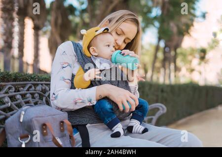 Bouteille d'alimentation de la mère-bébé garçon sur un banc de parc Banque D'Images