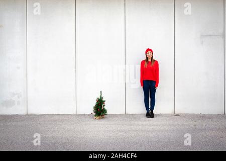 Woman wearing red pull et chapeau, wolly holding arbre de Noël artificiel devant un mur Banque D'Images