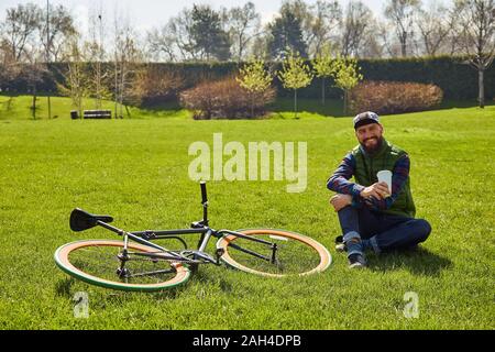 Jeune homme sur la pelouse avec un vélo. Il est titulaire d'un verre de café dans sa main. Dans la ville de loisirs Banque D'Images