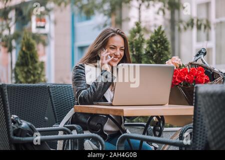 Portrait de belle jeune femme assise i street cafe avec ordinateur portable et parler au téléphone avec des amis Banque D'Images