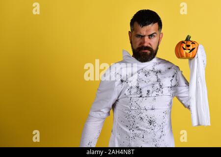 L'homme avec toute l'expression visage sur fond jaune, copiez l'espace. Heureux temps des Fêtes Halloween concept. Guy avec barbe est titulaire d'orange citrouille avec sourire. Caractère dans Halloween Costume fantôme blanc Banque D'Images