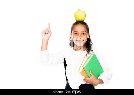 Cheerful african american lycéenne avec apple on head holding books et montrant idée geste isolated on white Banque D'Images