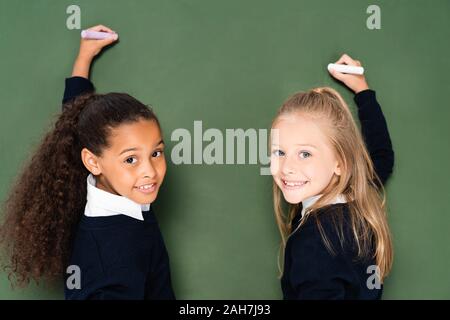 Deux écolières multiculturelle joyeuse smiling at camera while writing on chalkboard Banque D'Images