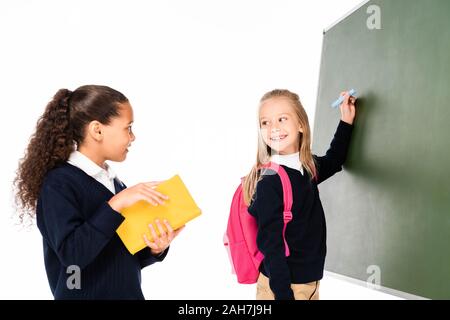 Deux smiling schoolgirl writing on clipboard et à la recherche à l'african american camarade holding book isolated on white Banque D'Images