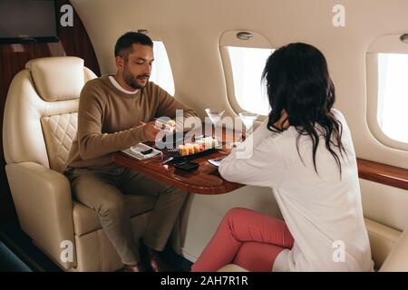 Vue arrière de femme assise avec l'homme près de sushi et verres en jet privé Banque D'Images