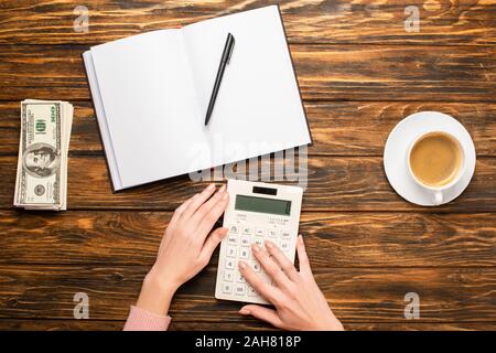 Portrait of businesswoman using calculator près de l'ordinateur portable blanc, billets de dollar et tasse de café sur un bureau en bois Banque D'Images