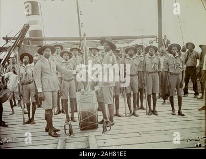 Le Nigeria, le marquage au verso : 'Scout Masters remise la troupe, Badagry, 1928'. Changer de montre ou de repos entre deux dirigeants des Boy Scouts du Nigeria à bord d'un bateau à vapeur. Les deux maîtres du Scoutisme se serrer la main dans un grand tambour électrique qui se dresse entre eux, regardé par un public d'au moins 20 African Boy Scouts qui sont tous debout à l'attention, 1928. 2/39/2000/098. Banque D'Images