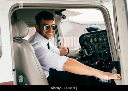 Smiling handsome pilote en lunettes looking at camera et showing thumb up en avion Banque D'Images
