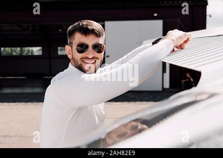 Homme barbu souriant à lunettes, debout près de avion en journée ensoleillée Banque D'Images