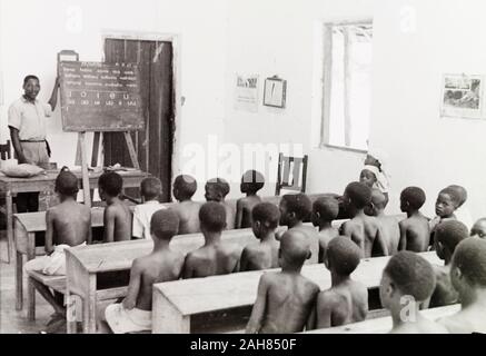 Au Kenya, les élèves assistent à une leçon de lecture dans une école au Kenya. Leur professeur se tient à côté d'un tableau noir inscrit avec voyelles et leurs associés à la phonétique, peut-être une indication qu'il s'agit d'une leçon d'anglais. Manuscrit original description : une école à la côte, 11 février 1947. 1995/076/1/2/7/31. Banque D'Images