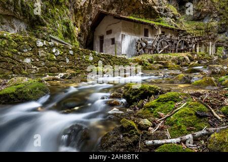 Vue à grand angle d'un ruisseau de montagne qui coule entre des rochers recouverts de mousse, avec un ancien moulin abandonné sur le fond Banque D'Images