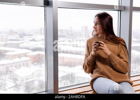 Jeune femme avec une tasse de café se trouve à côté de la fenêtre à l'étage supérieur d'un grand bâtiment, regardant vers le bas sur la ville par une journée d'hiver Banque D'Images