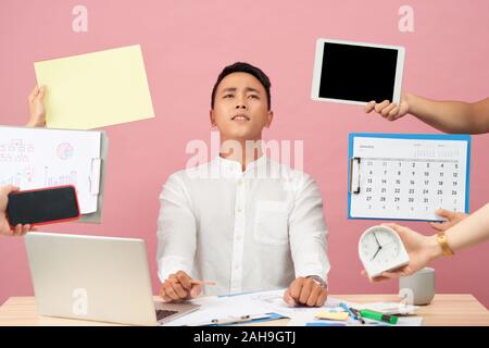 Jeune homme triste se trouve au bureau, mains avec des papiers, réveil, touchpad, le bloc-notes avec des autocollants, des études, de la documentation plus isolé sur fond rose Banque D'Images