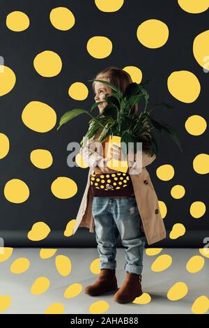 Cute child en jeans et trench-coat holding plant en pot sur fond noir avec des points jaunes abstract illustration Banque D'Images