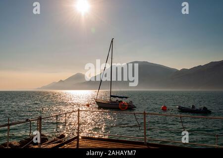Yacht à voile flottant près de la jetée sur le lac de Garda misty avec des dolomites avec soleil qui brille au-dessus dans le ciel sur l'arrière-plan Banque D'Images