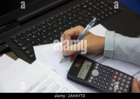 A woman's hand écrit avec un stylo sur un papier à côté d'un ordinateur portable, une calculatrice et plusieurs feuilles autour de Banque D'Images