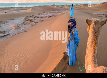 Désert du Sahara, au Maroc - le 25 octobre 2015. Une scène de désert à l'aube avec une tribu nomades portant des vêtements traditionnels, debout avec un chameau. Banque D'Images