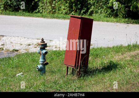 Se penchant dark red metal fire zone service verrouillé avec un cadenas à côté de rouillé délabré à incendie bleu entouré d'herbe non coupée Banque D'Images