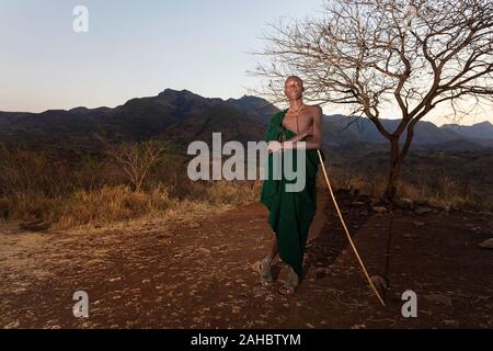 Jeune homme de la tribu Suri portant couverture verte debout sous un arbre et tenant un long bâton, de l'Ouest Banque d'Omo en Ethiopie Banque D'Images