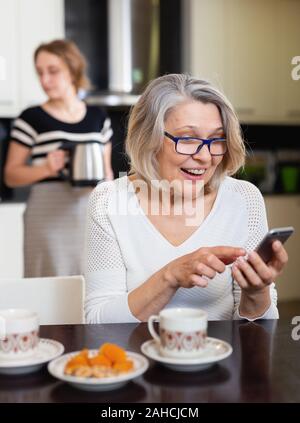 Couple positif à l'aide de téléphone pendant la jeune femme faire du thé Banque D'Images