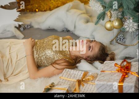 Jeune fille dans une robe de soirée. La fille est de rêver. La veille de Noël. Maison de vacances confortable au sapin avec des lumières et décoration dorée. fille dans une robe brillante sur Banque D'Images