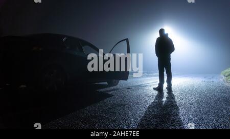 Un homme debout à côté d'une voiture, avec porte ouverte, garé sur le côté de la route, sous un lampadaire, sur un sinistre, effrayant, rural, route de campagne. Sur un Banque D'Images