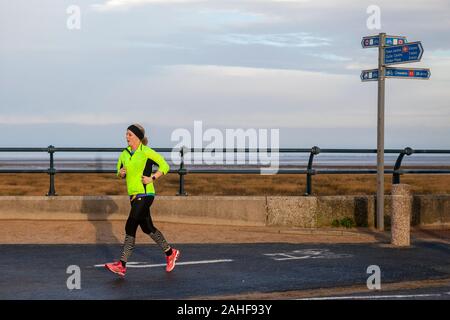 Southport, Merseyside, Royaume-Uni. 29 décembre 2019 Météo France. Des couleurs vives, des vêtements haute visibilité jaune, sur un fond gris pour la journée aussi sportive les résidents de prendre exercice tôt sur le front de la promenade côtière au nord-ouest de la station balnéaire. Credit : MediaWorldImages/Alamy Live News Banque D'Images