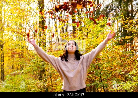 Jeune femme dans un chandail beige se trouve dans la forêt d'automne les feuilles et les jette en l'air Banque D'Images