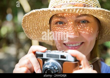 Gros plan portrait d'une belle femme en paille de chapeau de voyage dans la forêt tropicale, prenant des photos sur un appareil photo rétro. Ombres lumineuses grâce aux détails de découpe Banque D'Images