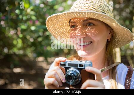 Gros plan portrait d'une belle femme en paille de chapeau de voyage dans la forêt tropicale, prenant des photos sur un appareil photo rétro. Ombres lumineuses grâce aux détails de découpe Banque D'Images