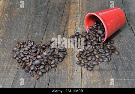 Café en grains en forme de coeur à côté d'un rouge renversé tasse pleine de haricots sur fond de bois Banque D'Images
