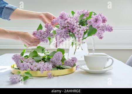 Mains de Femme avec bouquet de fleurs lilas en verseuse en verre, l'arrière-plan de table blanc, tasse de thé. Printemps à la décoration d'intérieur avec des fleurs Banque D'Images