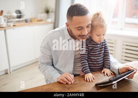 Portrait chaleureux de père aimant avec cute little girl using digital tablet, tandis qu'assis à table en bois dans la cuisine confortable, copy space Banque D'Images