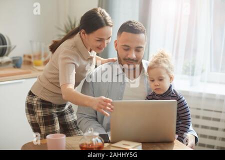 Portrait chaleureux de race mixte moderne-famille à l'aide d'ordinateur tout en étant assis dans de confortables cuisine intérieur avec mignon fille Banque D'Images