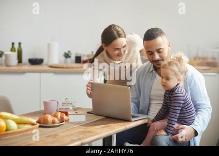 Portrait chaleureux de race mixte moderne-famille à l'aide d'ordinateur tout en étant assis dans de confortables cuisine intérieur avec mignon fille, copy space Banque D'Images