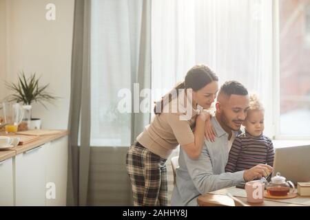 Portrait aux teintes chaleureuses de race mixte moderne-family using laptop while sitting in cuisine confortable intérieur avec jolie petite fille, copy space Banque D'Images