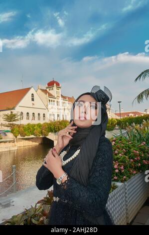 Jeune femme portant le hijab noir sur un après-midi ensoleillé de regarder le ciel portant un collier de perle debout à côté d'une rivière Banque D'Images