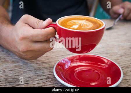 Tenir la main de l'homme red tasse de café avec du lait pour faire de photo de fleurs dans l'assiette et cuillère puis le soulever sur table en bois écorce Banque D'Images