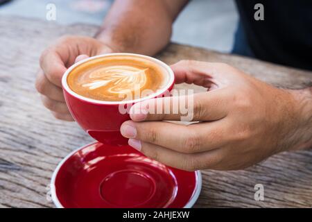 Tenir la main de l'homme red tasse de café avec du lait pour faire de photo de fleurs dans l'assiette et cuillère puis le soulever à deux mains sur la table en bois écorce Banque D'Images